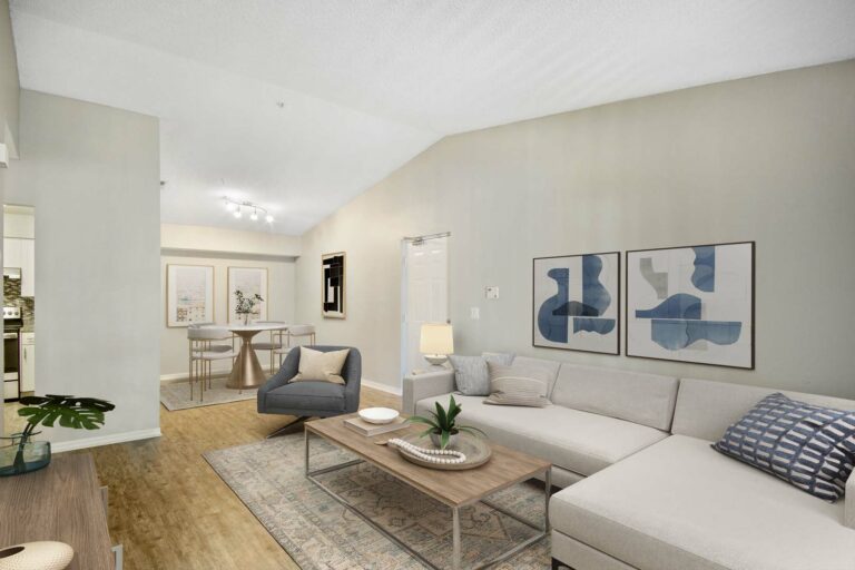 Virtually staged living room with vaulted ceilings hardwood-style flooring, couch, decorative rug, and coffee table looking into the dining room with table and chairs.