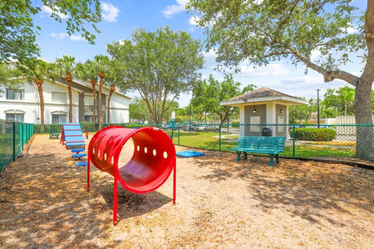Fenced in dog park with agility course, benches, and apartments in the background surrounded by native landscaping.